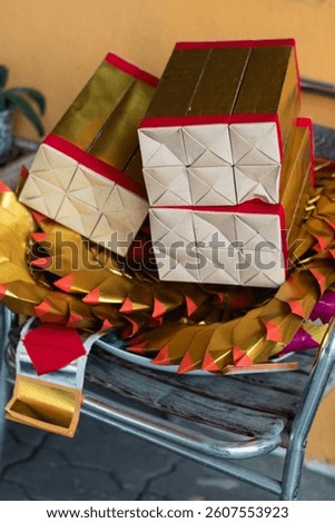 A photo shows stacks of joss paper, likely for burning during the Qingming Festival. The gold and silver paper, some folded into ingot shapes, rests on a chair against a yellow wall.
