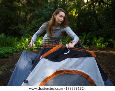 The photo shows a sunny morning in the forest. A young woman in a campground is laying out a tent. The woman has long blonde hair and sportswear. In the background is a green forest.