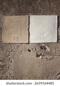 The photo shows two plaster or rectangular tile samples side by side on a rough, textured wall, presumably masonry or stucco. The sample on the left is beige or tan in color and has the number 