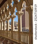 The photo shows a Venetian Gothic-style colonnade with ornate arches and carved capitals. Sunlight highlights the stone