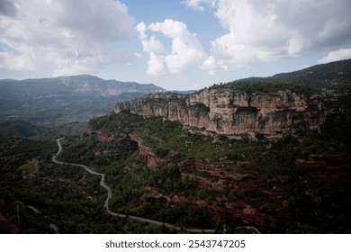 The photo shows a winding road cutting through a lush, green valley surrounded by steep cliffs. The landscape is dominated by rugged rock formations and distant mountains in Siurana, in Spain. - Powered by Shutterstock