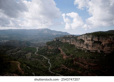 The photo shows a winding road cutting through a lush, green valley surrounded by steep cliffs. The landscape is dominated by rugged rock formations and distant mountains in Siurana, in Spain. - Powered by Shutterstock