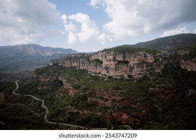 The photo shows a winding road cutting through a lush, green valley surrounded by steep cliffs. The landscape is dominated by rugged rock formations and distant mountains in Siurana, in Spain. - Powered by Shutterstock