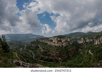 The photo shows a winding road cutting through a lush, green valley surrounded by steep cliffs. The landscape is dominated by rugged rock formations and distant mountains in Siurana, in Spain. - Powered by Shutterstock