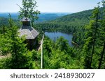 The photo shows a wooden chapel perched on a rocky outcrop surrounded by dense forest above a mountain lake. Taken at the Rachel Chapel in the Bavarian Forest National Park, Germany.