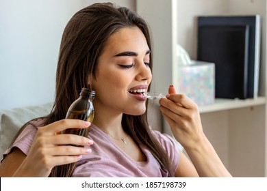 Photo Of A Sick Young Woman Sitting On The Bed Of The House And Drink The Bitter Medicine. Woman Taking Cough Syrup On Sofa At Home. Young Woman Pouring Cough Syrup. Health, Flu And People Concept.