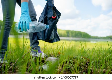 Photo of side of girl in rubber gloves picking up garbage in bag on banks of river.
