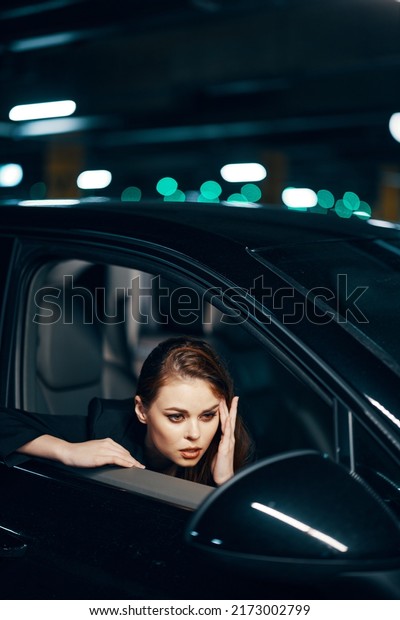 photo from the side, at night, of a woman sitting in\
a black car and looking out of the window and gently touching her\
face looks into the side view mirror after parking the car in the\
parking lot