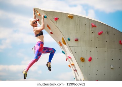 Photo from side of sports woman in leggings hanging on wall for climbing against blue sky