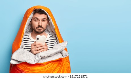 Photo Of Sleepy Unshaven Man Has Tired Sleepy Expression, Wakes Up Early In Morning, Spends Night In Open Air, Holds Modern Mobile Phone, Sends Text Messages, Poses Over Blue Background. Camping