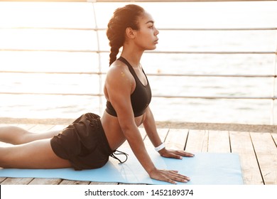 Photo of slim woman wearing sportive clothes stretching her body on finess mat while doing workout by seaside in morning