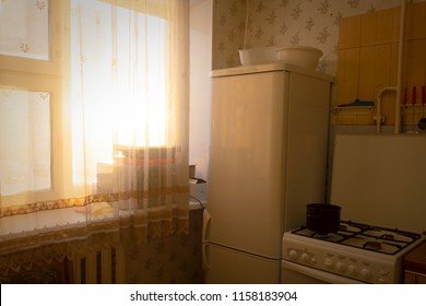 Photo Of A Small Cozy Kitchen. Sunset In The Kitchen. Sunset Through The Window. Warm Orange Light. Refrigerator And Stove In The Small Kitchen. Old Interior