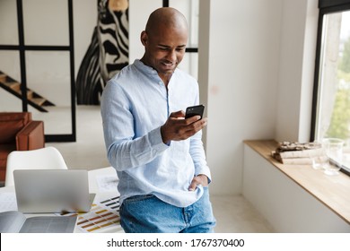 Photo of smiling african american man using mobile phone while working in living room at home - Powered by Shutterstock