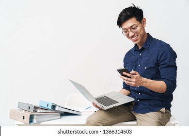 Photo of smiling asian man 20s wearing eyeglasses holding smartphone and laptop while working in office isolated over white background