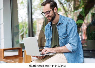 Photo of smiling bearded man wearing glasses typing on laptop while working in cafe outdoors - Shutterstock ID 1414673465