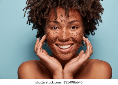 Photo of smiling black woman has cleansing mask or scrub on face, smiles broadly, enjoys beauty treatments in salon, touches soft skin, looks happily at camera, isolated over blue background