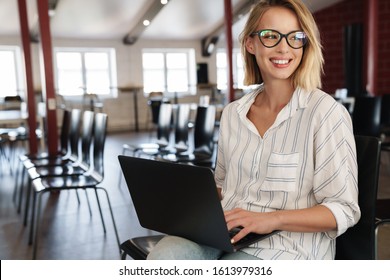 Photo of smiling blonde woman wearing eyeglasses working on laptop while sitting in conference hall
