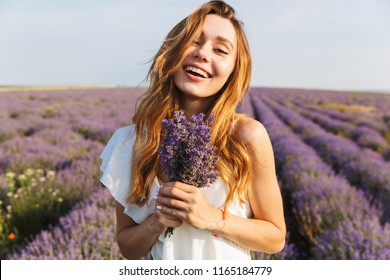 Photo of smiling brunette woman in dress holding bouquet with flowers while walking outdoor through lavender field in summer