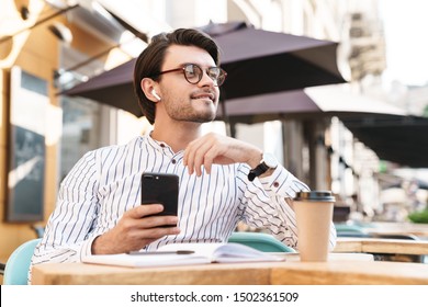 Photo of smiling caucasian man wearing eyeglasses using earpod and cellphone while working in cafe outdoors - Shutterstock ID 1502361509