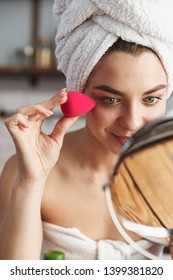 Photo of smiling caucasian woman wrapped in white towel applying makeup with cosmetic sponge in apartment
