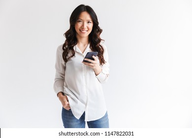 Photo of smiling charming asian woman with long dark hair holding black cell phone isolated over white background in studio