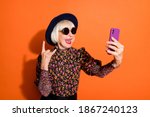 Photo of smiling cheerful happy modern grandmother take selfie on phone show rock n roll sign isolated on orange color background