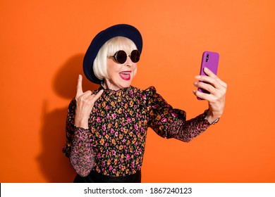 Photo of smiling cheerful happy modern grandmother take selfie on phone show rock n roll sign isolated on orange color background