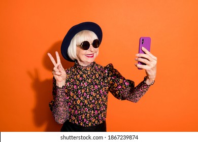 Photo of smiling cheerful positive good mood old woman take selfie on phone showing v-sign isolated on orange color background - Powered by Shutterstock