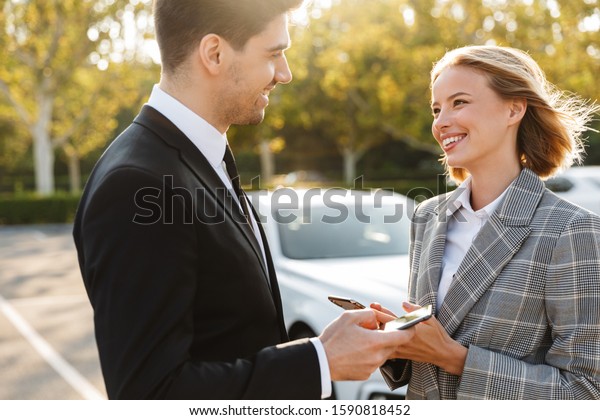 Photo of smiling\
colleagues man and woman in formal wear talking and using\
cellphones on parking\
outdoors