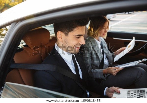 Photo of smiling\
colleagues man and woman in formal wear working with documents and\
laptop while going in car