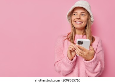 Photo of smiling European woman looks with happy expression uses mobile phone for chatting online wears winter panama and cashmere jumper isolated over pink background copy space for promotion
