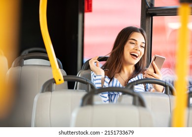 Photo Of Smiling Girl Sitting In Bus. Female Is Using Mobile Phone While Commuting By Public Transport. She Is Wearing Blue Shirt.