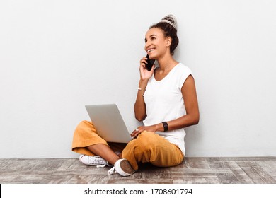 Photo of a smiling happy young african woman with dreads sitting isolated over white wall background using laptop computer talking by mobile phone. - Powered by Shutterstock