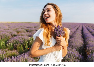 Photo of smiling joyful woman in dress holding bouquet with flowers while walking outdoor through lavender field in summer