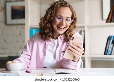 Photo of smiling nice woman wearing eyeglasses using cellphone and drinking coffee while doing homework in room - Powered by Shutterstock