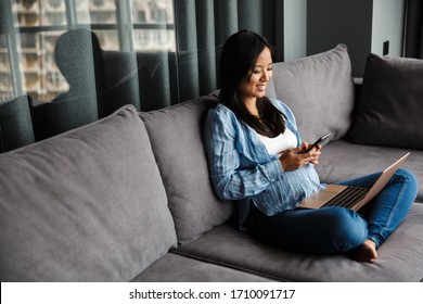 Photo Of Smiling Pregnant Asian Woman Using Laptop And Mobile Phone While Sitting On Couch At Home