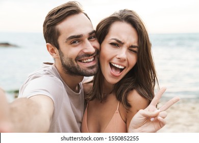 Photo of smiling young couple gesturing peace sign and taking selfie photo while resting on sunny beach