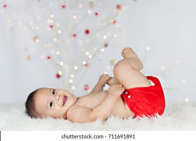 Photo Of A Smiling,healthy, Chubby Baby Lying On His Back On A White Sheepskin, Wearing A Red Cloth Diaper/nappy With A Lit, White Christmas Tree In The Background.