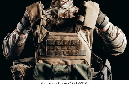 Photo of soldier in camouflaged uniform and tactical gloves holding military armored vest on black background.