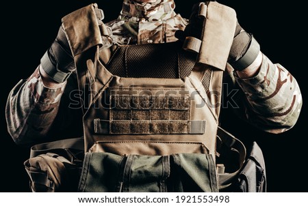 Photo of soldier in camouflaged uniform and tactical gloves holding military armored vest on black background.