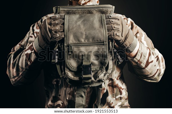Photo of soldier\
in camouflaged uniform and tactical gloves holding leg bag on black\
background close-up view.