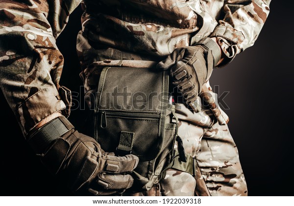 Photo of soldier in camouflaged\
uniform and tactical gloves using leg bag on black\
background.