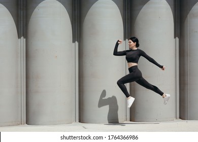 Photo Of A Sports Asian Woman In A Tight-fitting Suit Who Froze Sideways In A Jump On The Street Against The Background Of Concrete Walls