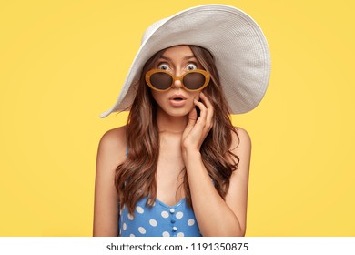Photo of startled young Caicasian lady stares with bugged eyes, wears trendy sunglasses, summer hat, recieves unexpected news, poses over yellow background. People, emotions and reaction concept