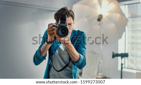 In the Photo Studio with Professional Equipment: Portrait of the Famous Photographer Holding State of the Art Camera Taking Pictures with Softboxes Flashing in Background.