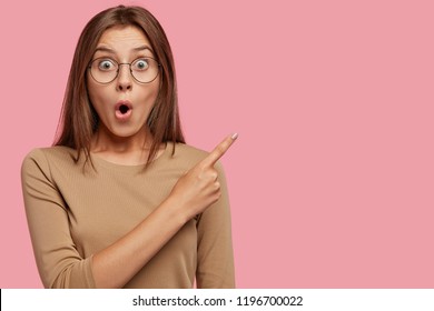 Photo of stupefiied Caucasian girl stares at camera, keeps jaw dropped, points with index finger at upper right corner, shows free space for your promotional content. Omg, look at this thing