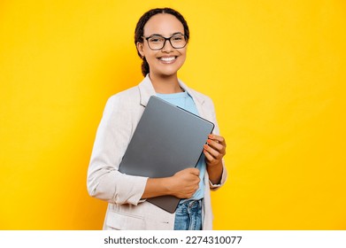 Photo of a successful confident brazilian or hispanic business lady in stylish elegant clothes, creative manager, holding laptop, stand on isolated yellow background, looks at camera, smiles friendly - Powered by Shutterstock