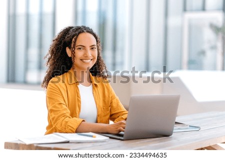 Photo of a successful curly haired girl, brazilian or hispanic nationality, outsourcing employee or student, uses laptop to work or study online while sitting outdoors, looks at camera, smile friendly