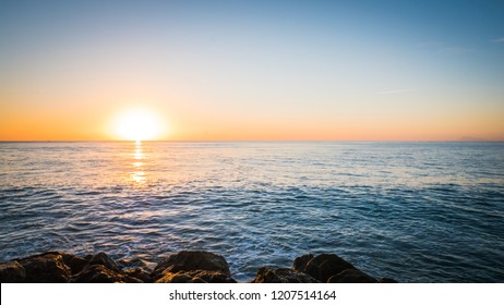 A photo of the sun rising over the Mediterranean Sea. The calm water reflects the sunlight like a mirror. The photo was taken in Punta Negra in the seaside town of Cullera, in Valencia, Spain, Europe 
