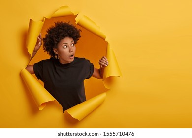 Photo of surprised Afro woman in black t shirt gasps from amazement, looks with scared face expression aside, dressed in black t shirt, unexpected to see something horrible, poses in torn paper wall